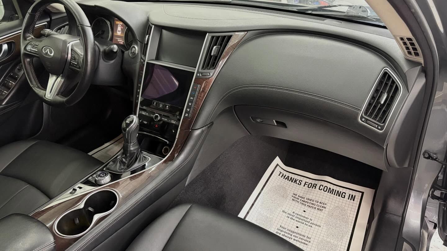 Interior view of a gray car dashboard with a black leather steering wheel, center console, and paper floor mat.