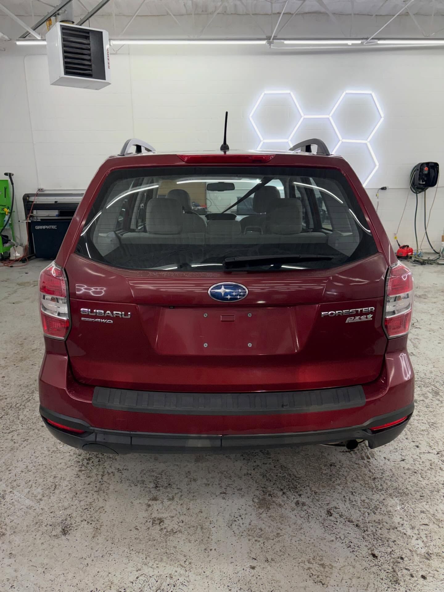 A red Subaru Forester seen from the rear, parked inside a brightly lit automotive workshop with a hexagonal light design.