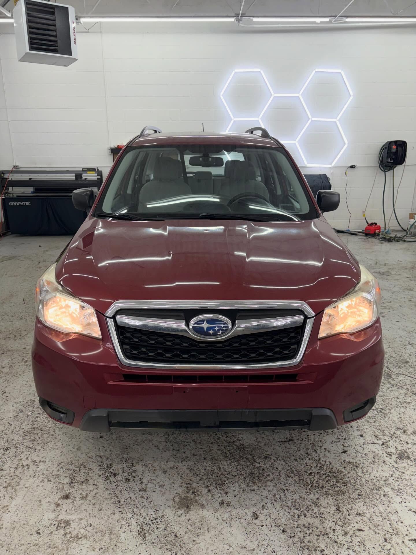 A front-facing view of a maroon Subaru Forester parked inside a professional garage with hexagonal wall lighting.