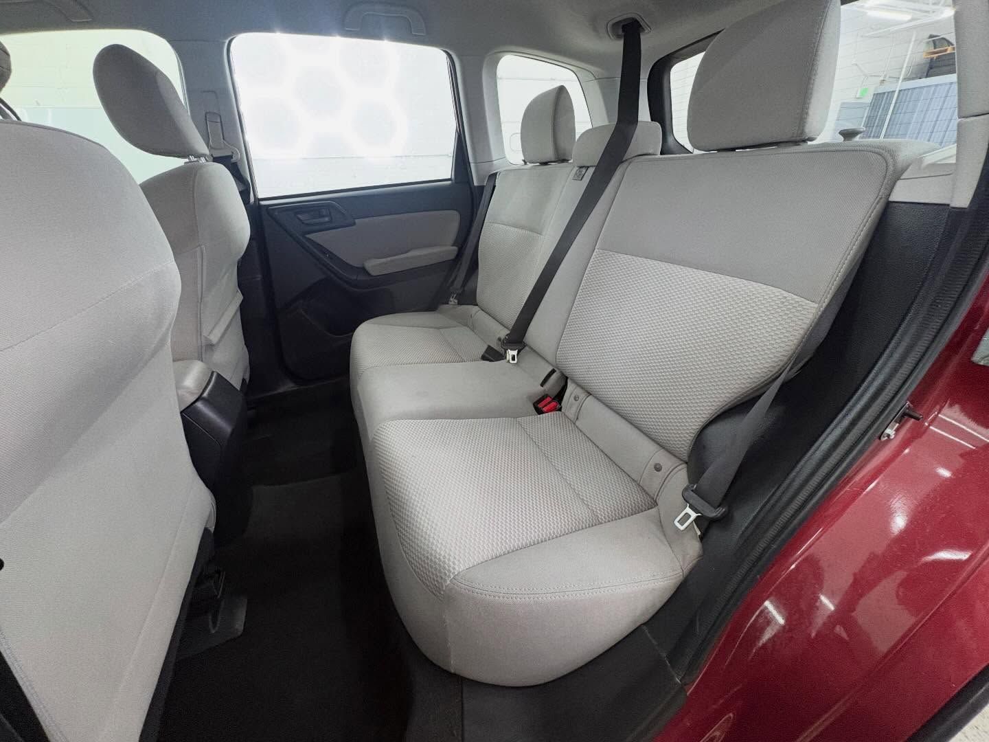 The interior back seat of a maroon vehicle, featuring light gray fabric upholstery, black floor mats, and seatbelts.