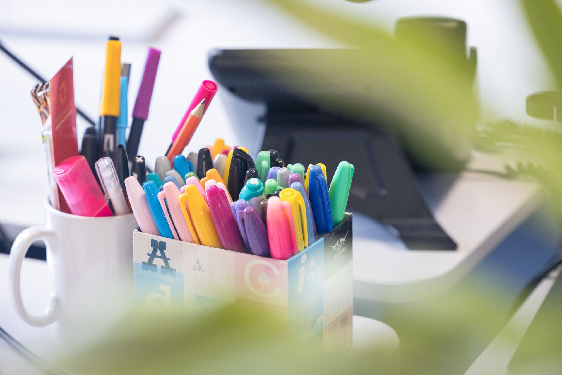 Colorful pens and markers in a white mug on a desk, with a blurred laptop in the background