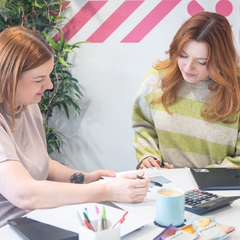 Two people collaborate at a table, reviewing papers with a calculator and markers in a bright office.