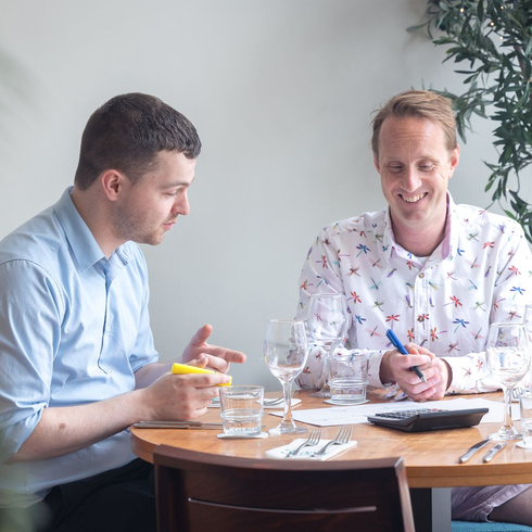 Two coworkers discussing papers at a table, one pointing with a yellow pen and the other smiling.