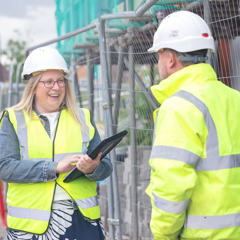 Two construction workers in hard hats and neon safety vests reviewing a tablet on site