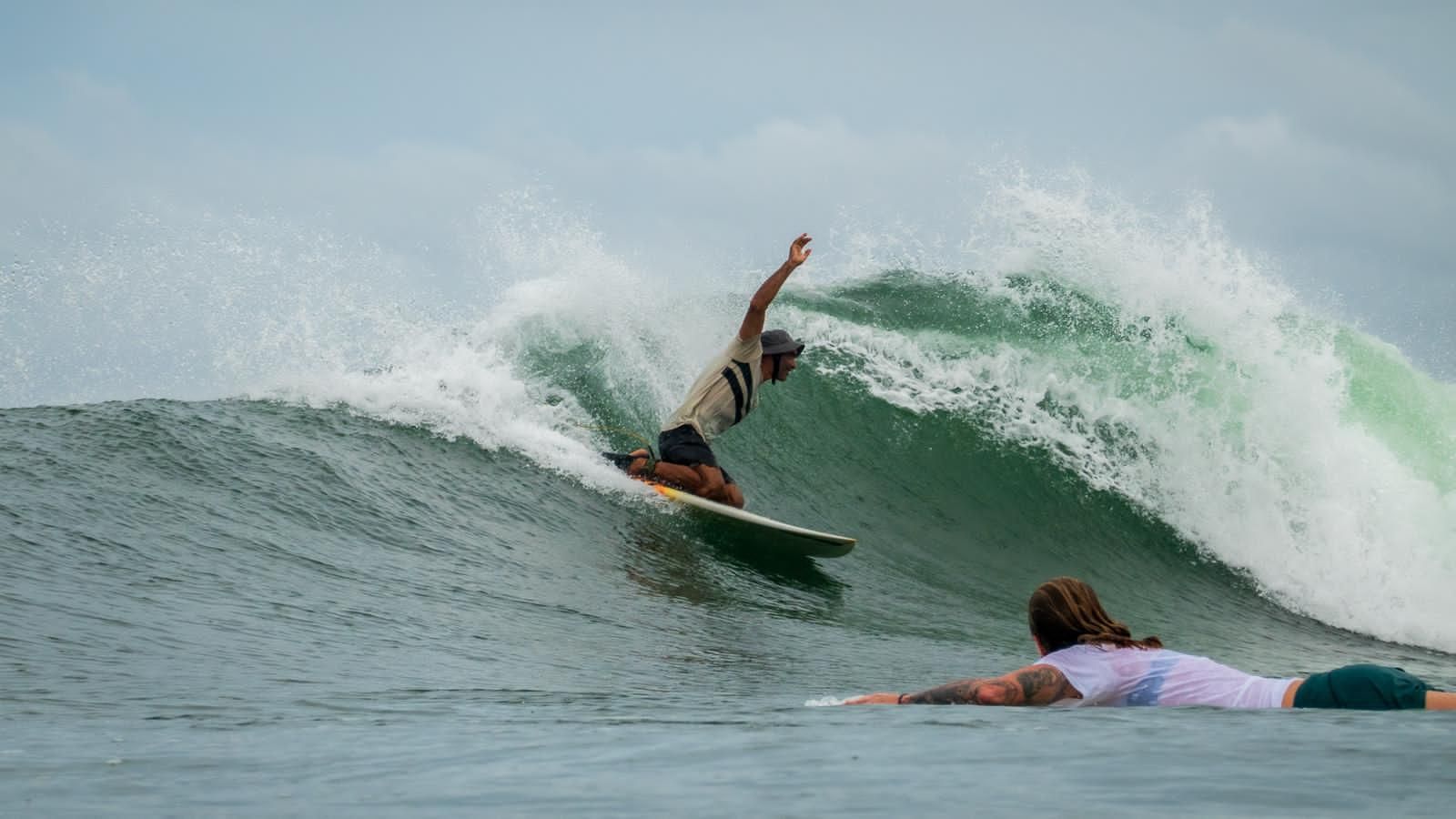 Surfer carving a wave, arm raised. Another surfer waits in the water. Cloudy day, green and white wave.