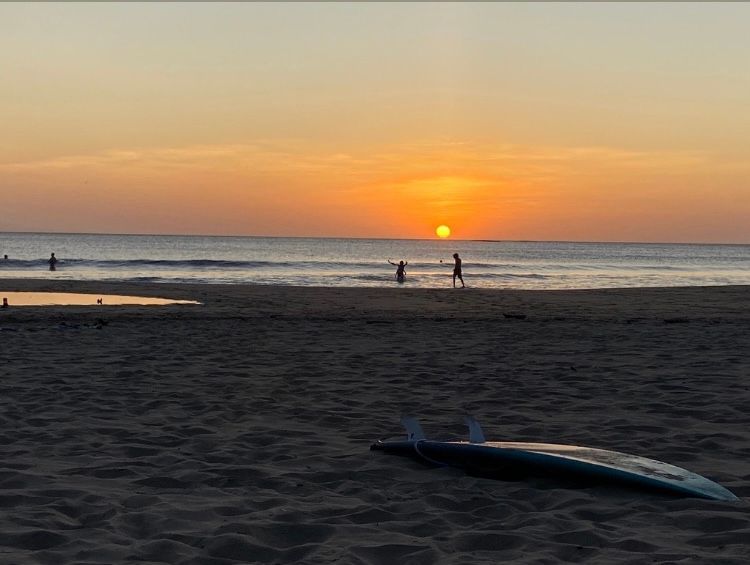 Sunset over ocean; person surfing at beach. Surfboard in foreground.