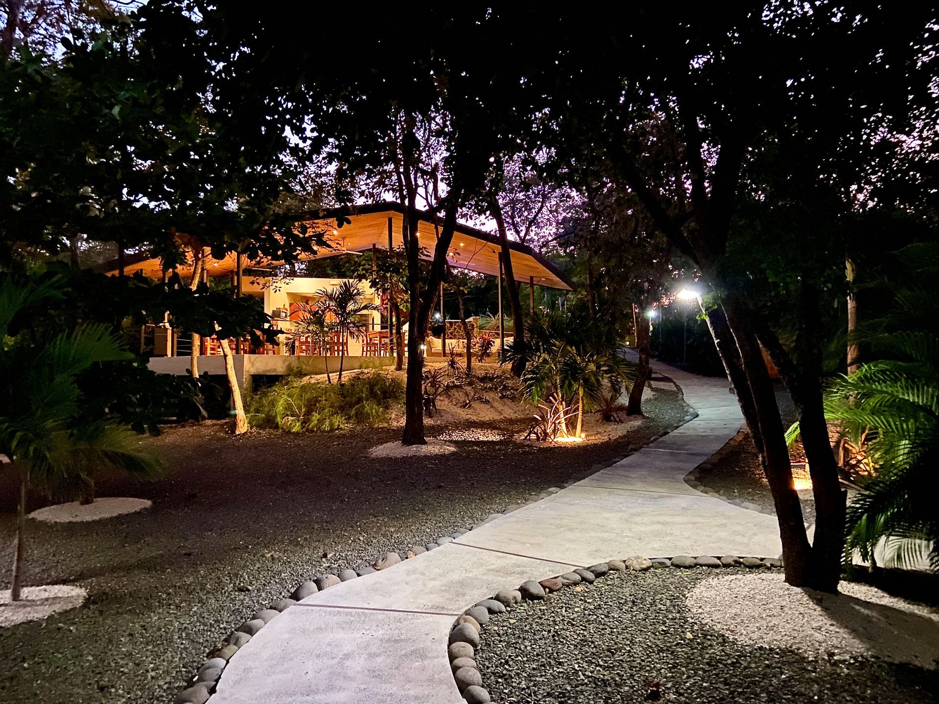 Stone path leads to a building at dusk, lit by outdoor lights. Trees surround the path and building.
