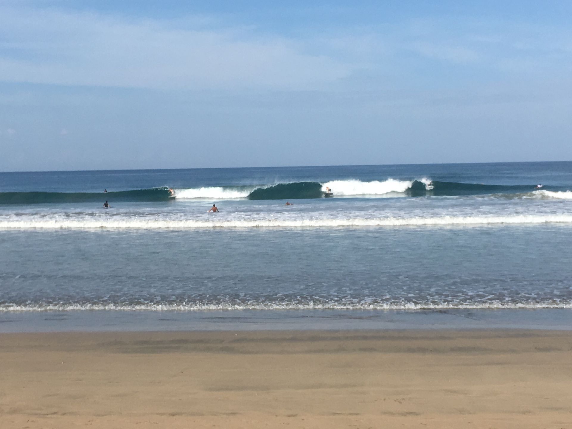 Waves breaking on a sandy beach; surfers in the water under a clear blue sky.