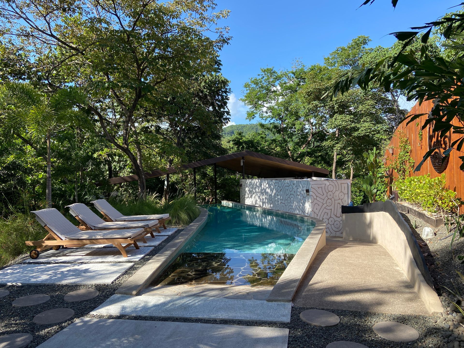Pool with lounge chairs surrounded by trees and a blue sky.