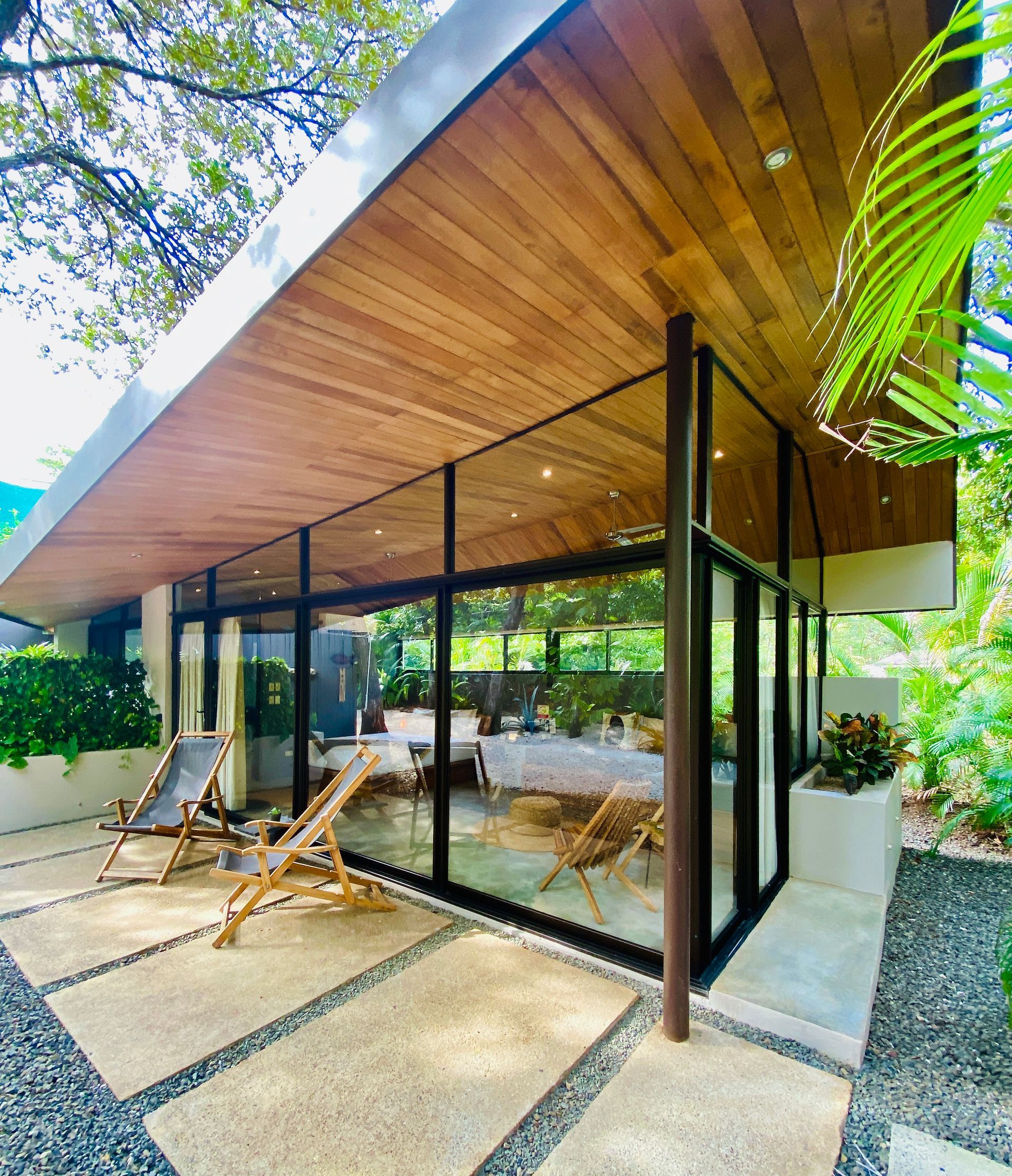 Modern home with wooden ceiling, large windows, and patio with lounge chairs surrounded by greenery.