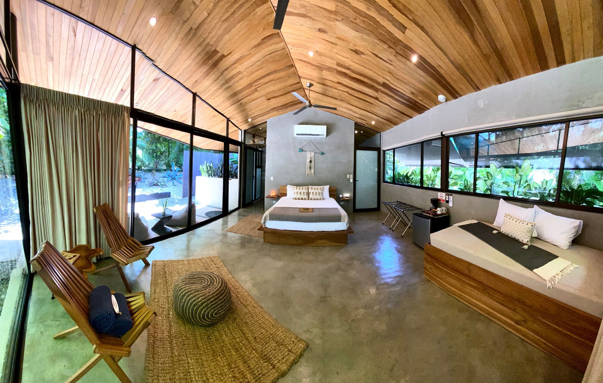 Spacious modern bedroom with glass walls, wooden ceiling, bed, seating, and surrounding greenery.
