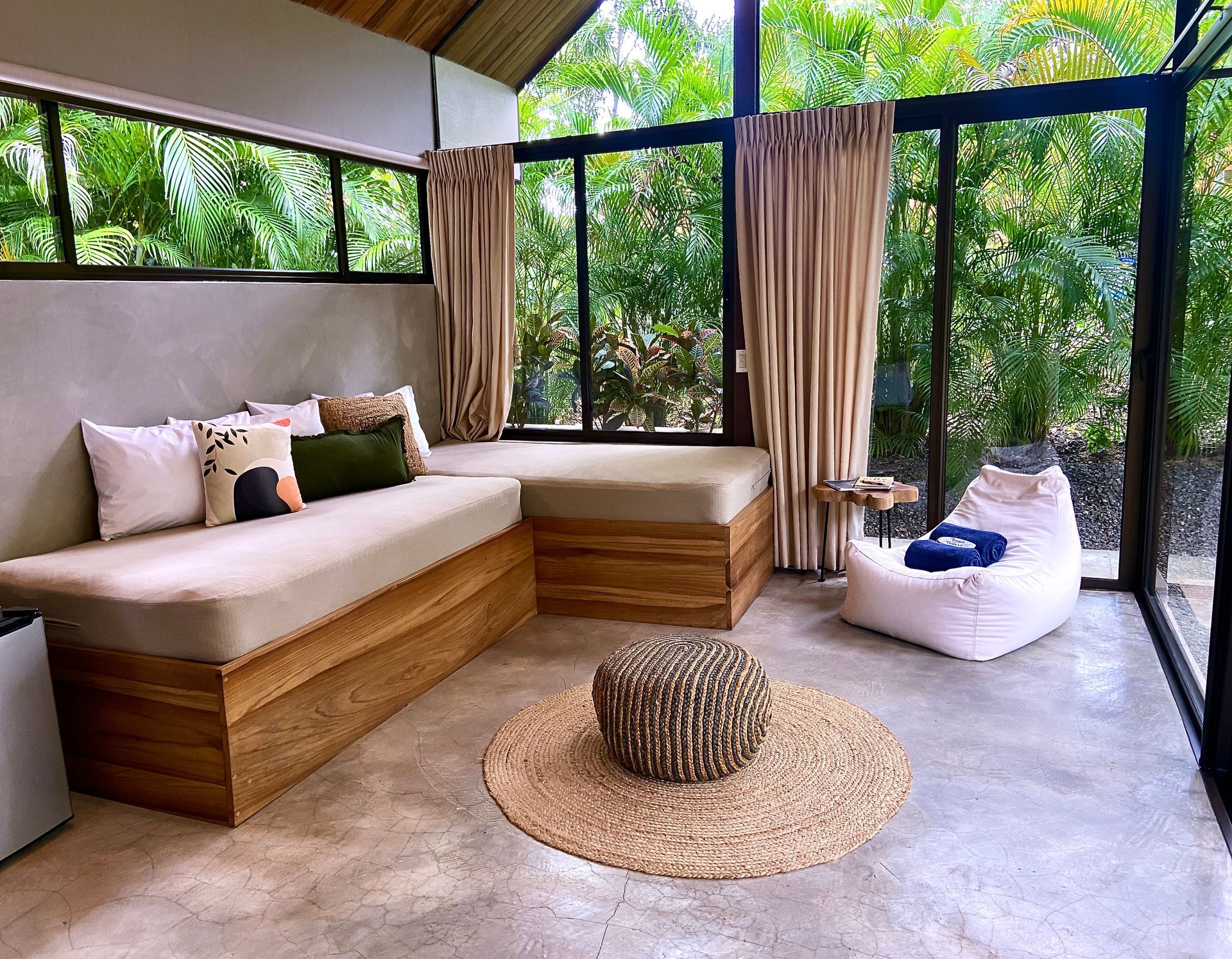 Cozy outdoor living room with a beige sectional, beanbag, and woven rug, surrounded by glass windows and lush greenery.