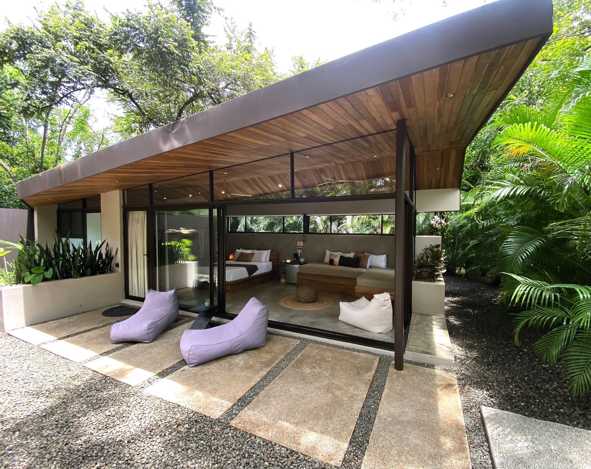Modern bungalow with light wood ceiling, glass walls, and purple bean bag chairs on a stone patio.