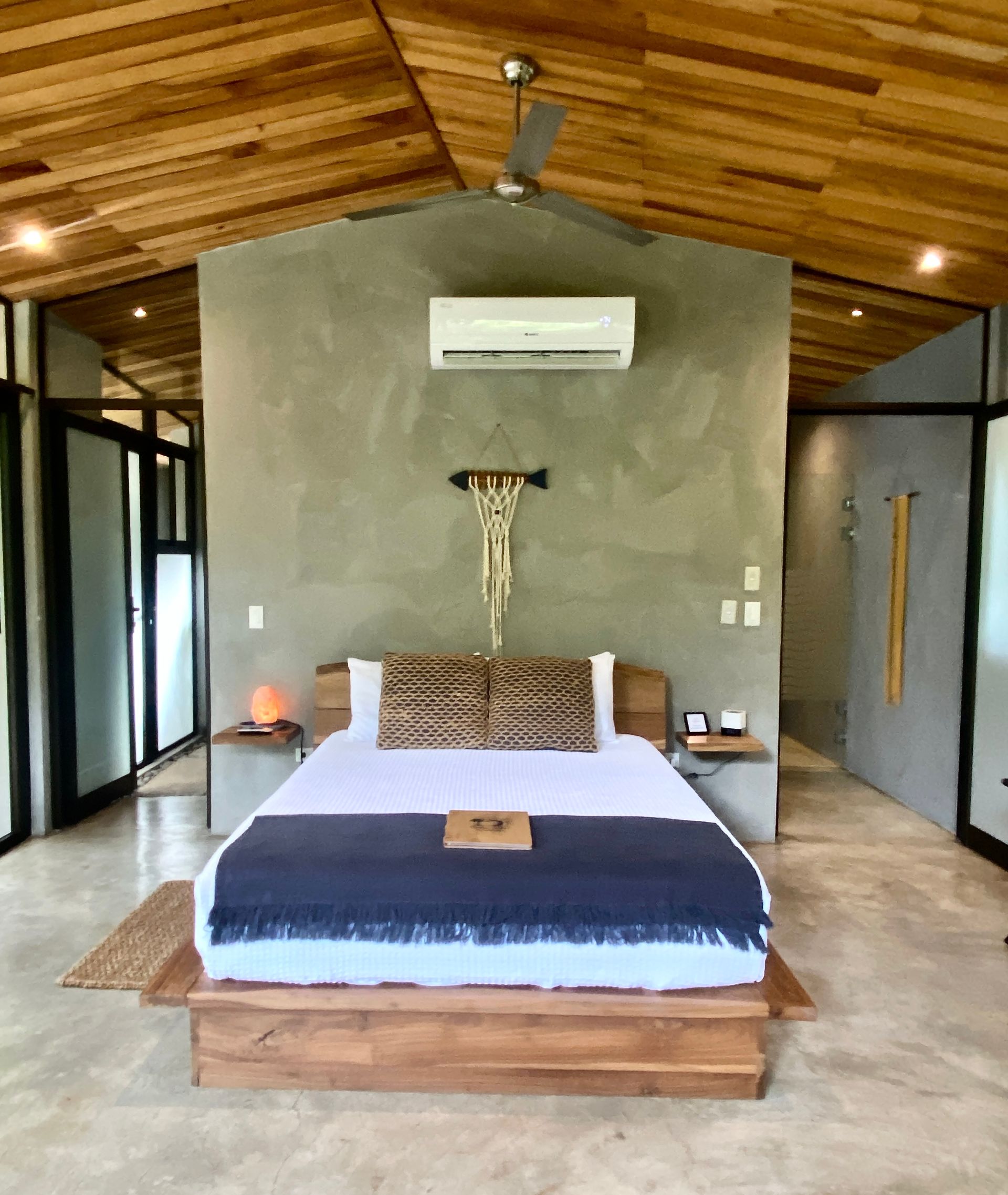 Bedroom with wooden ceiling and bed, gray wall with macrame, and floor-to-ceiling glass walls.
