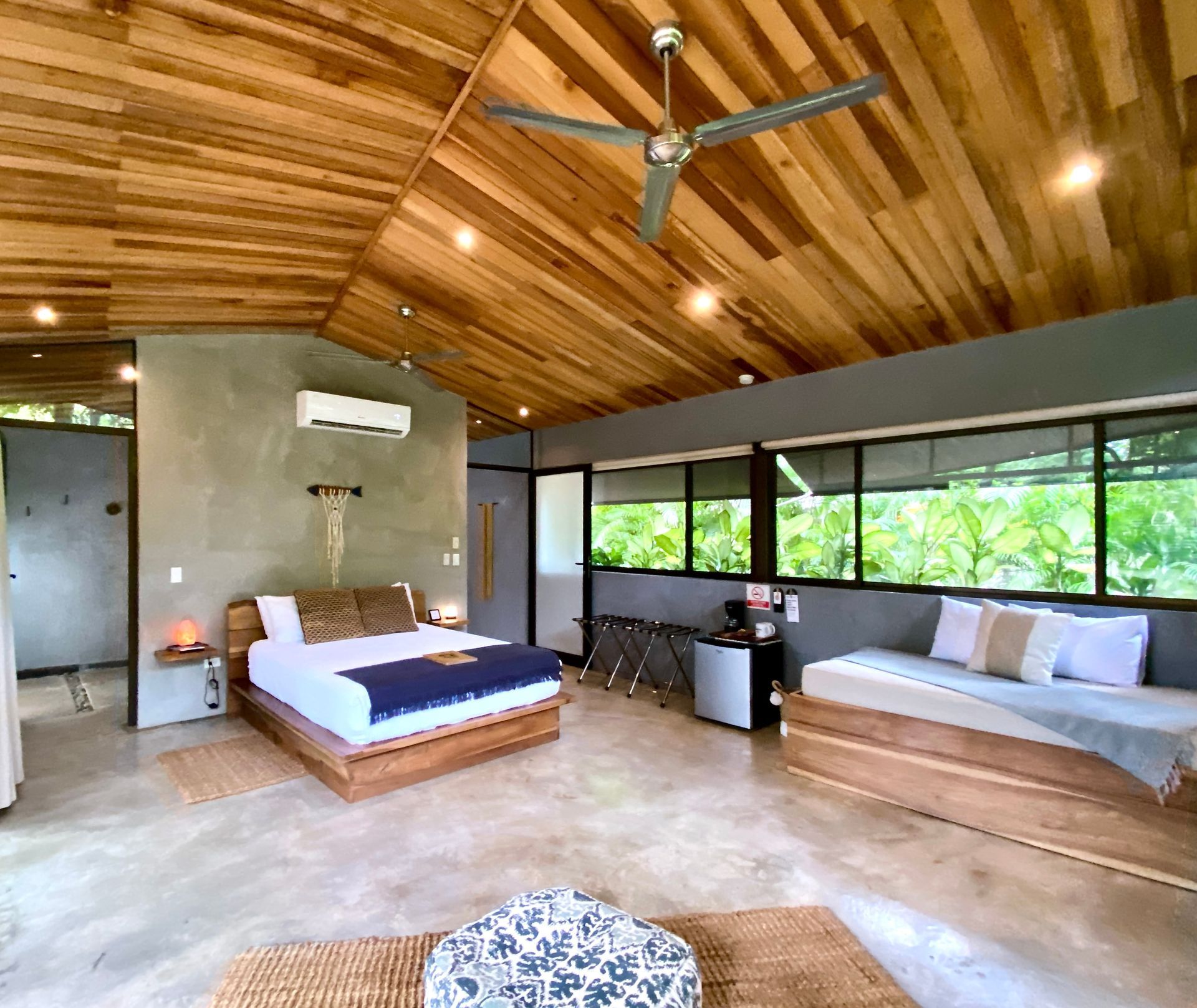 Bedroom with wooden ceiling and furniture, concrete floors, and large windows overlooking greenery.