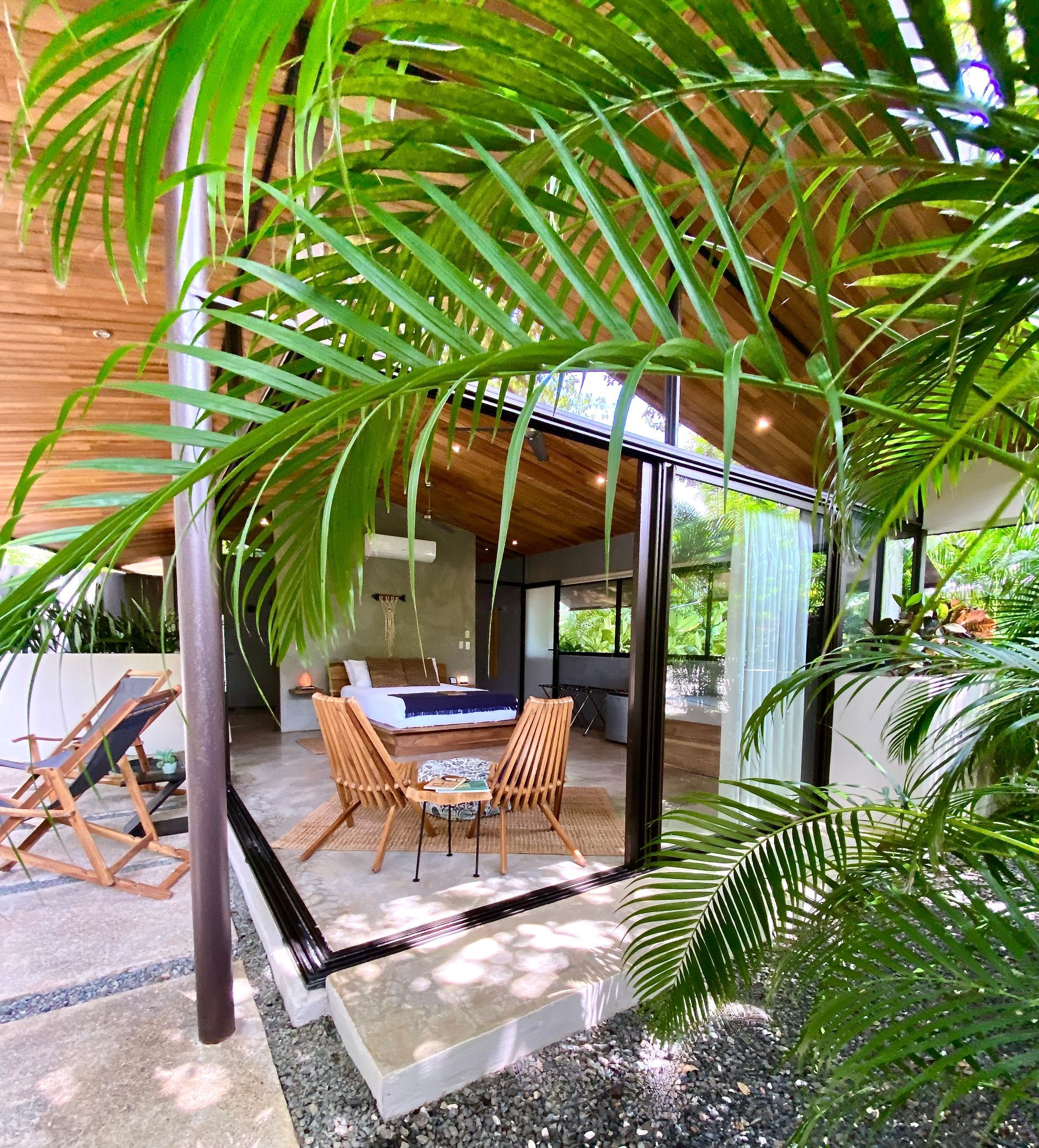 Open-air living space with wooden ceiling, stone floor, and tropical foliage. Furnishings include chairs and a small table.