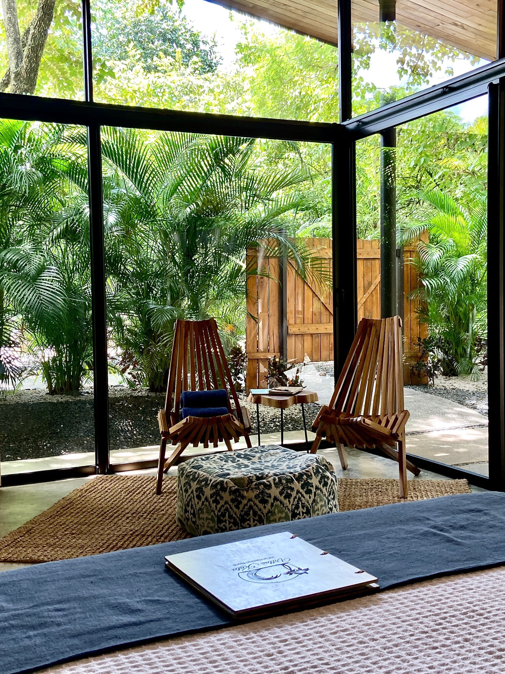 Two chairs, a small table, and a patterned ottoman sit in front of a wall of windows with outdoor foliage visible.