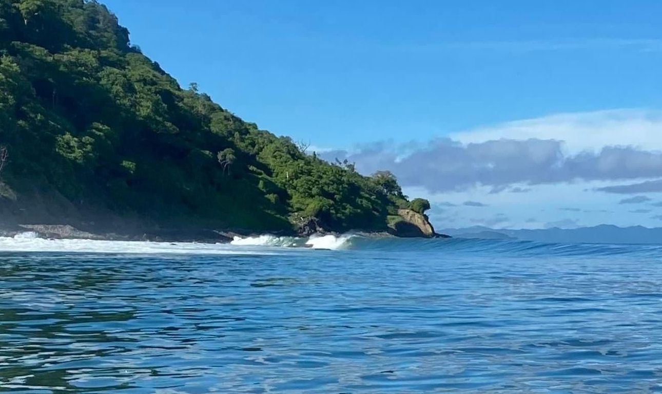 Ocean waves crashing near a lush, green hillside under a bright blue sky.
