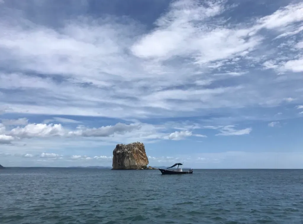 Ocean view: rocky island and small boat under a blue sky with scattered clouds.