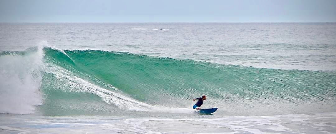 A surfer rides a teal wave, the ocean stretches out under a pale blue sky.