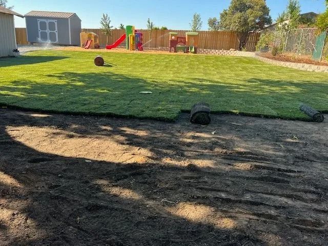 A backyard under renovation with newly laid sod on the upper lawn and unpaved dirt in the foreground.