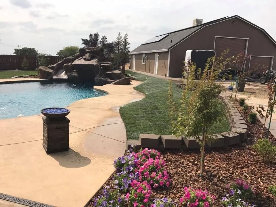 A backyard pool with a rock water feature, a curved grass lawn, flower beds, and a large barn-style building in the rear.