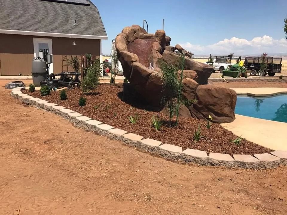 A large brown rock-shaped pool slide sits next to a pool in a yard with fresh mulch, retaining wall blocks, and a shed.
