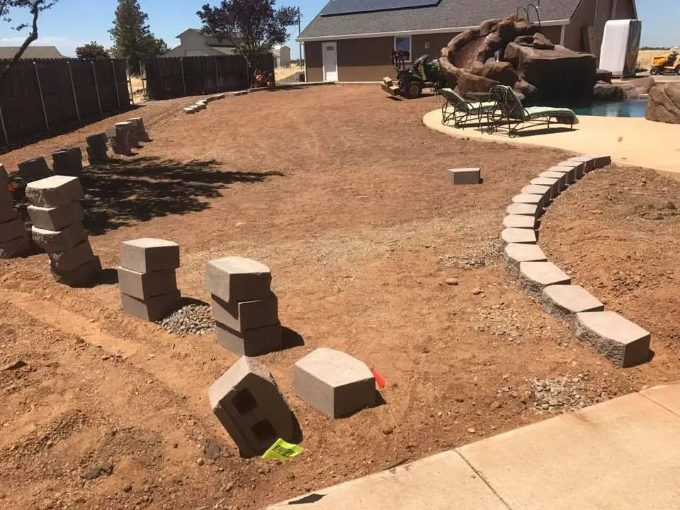 A residential backyard under construction with tan dirt, stacks of landscaping blocks, and a curving stone border.