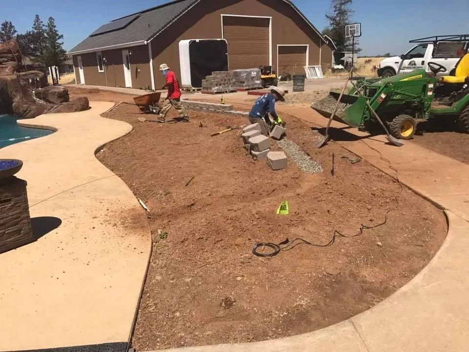 Two workers install a stone retaining wall in a dirt yard next to a concrete pool deck and a green tractor.