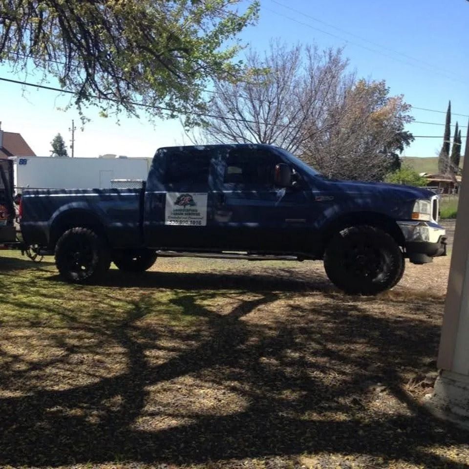A dark blue pickup truck with a business sign on the door parked on a grassy lot under trees.