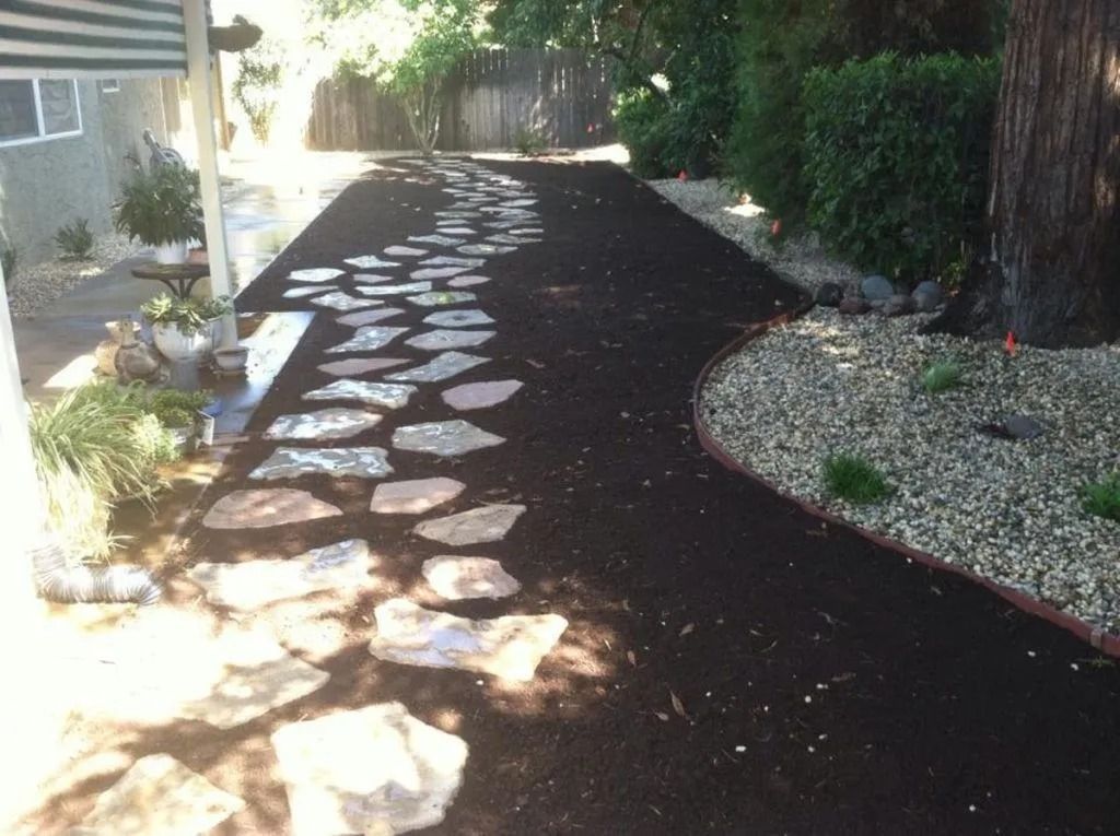 A stone stepping-stone path winds through a dark mulch yard beside a residential building and a rock garden.