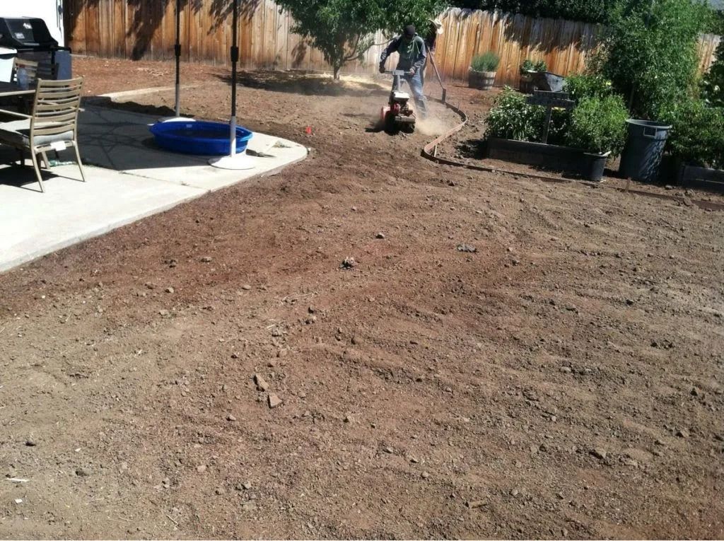 A person uses a rototiller to churn soil in a large, dirt-covered backyard.