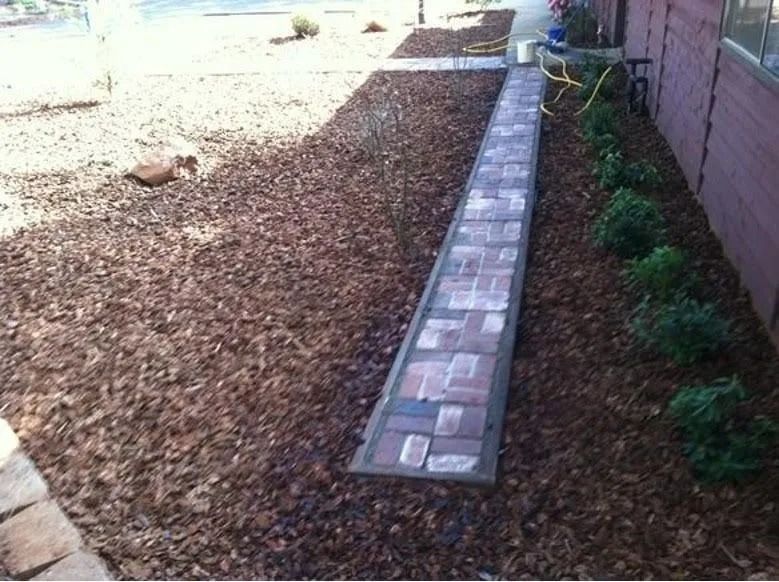 A narrow brick walkway cuts through a yard with brown mulch, positioned next to a brick building wall and small shrubs.