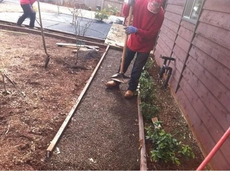 A person in a red shirt uses a shovel to spread dark mulch along a narrow pathway next to a wooden building.