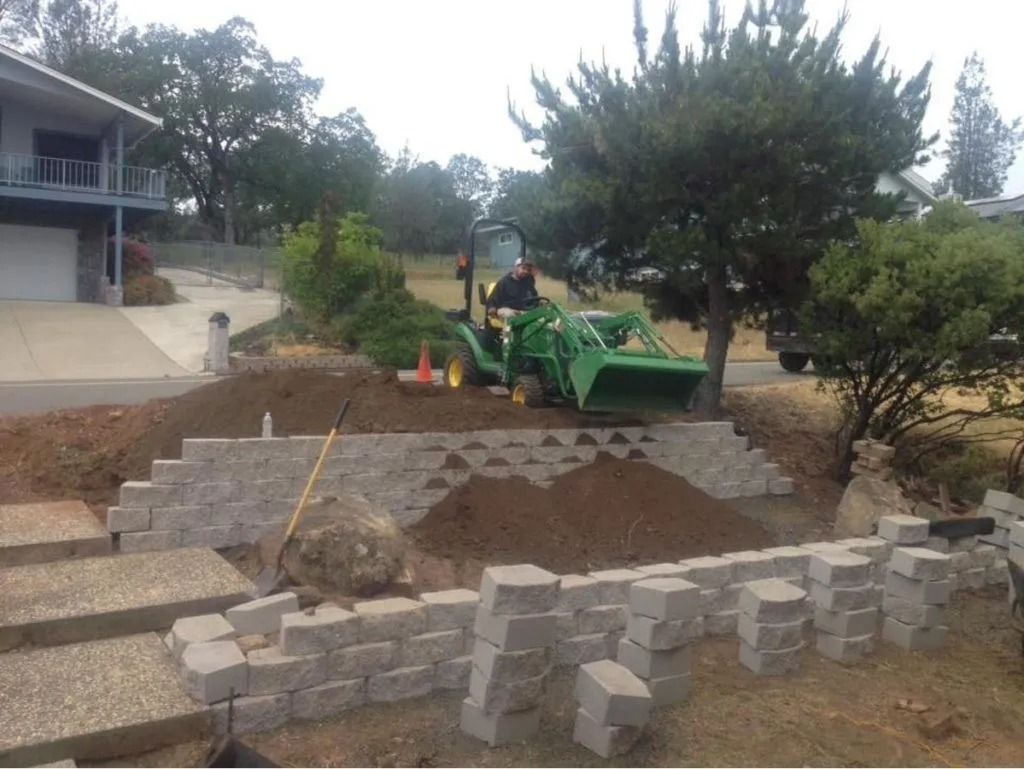 A green tractor works on a terraced residential yard featuring newly built retaining walls made of grey stone blocks.