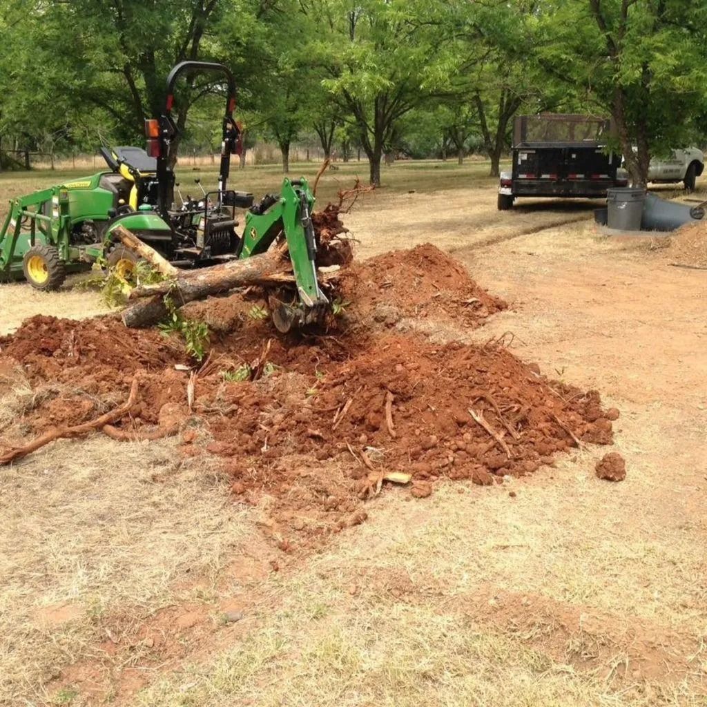A green tractor uses a backhoe attachment to remove a tree stump in a field of red soil near a trailer.