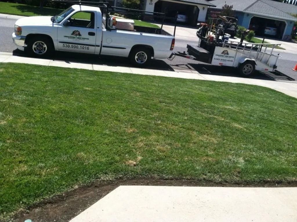 A white pickup truck with a logo on its side is parked on a suburban street, towing a small flatbed utility trailer.