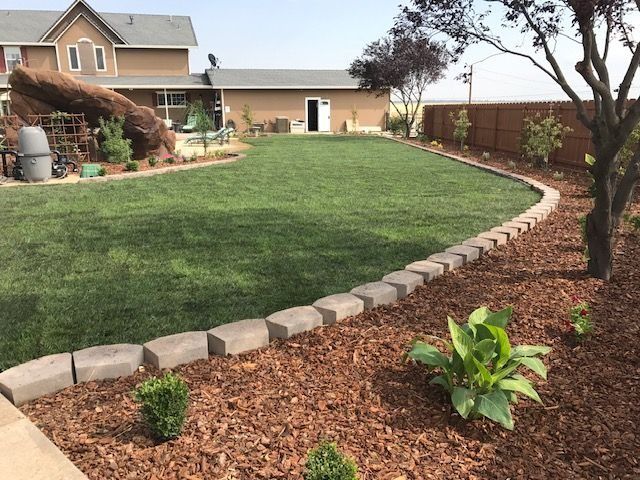 A landscaped backyard with a green lawn, curved stone border, mulch beds, and a large decorative rock feature.