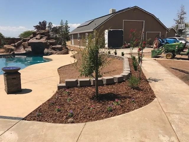 A backyard landscape featuring a pool with a rock waterfall, a retaining wall, mulch garden beds, and a large barn.