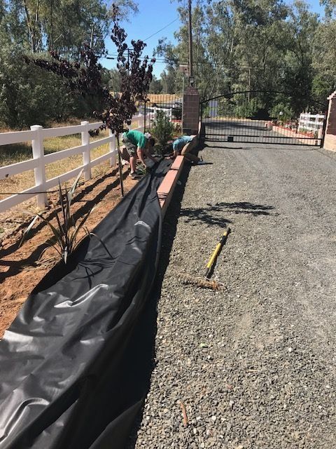 Two people installing landscape fabric along a white fence next to a gravel driveway.