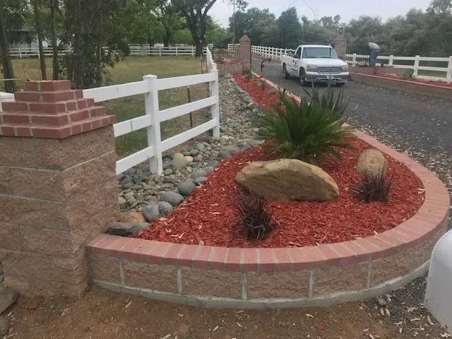 A landscaped garden bed with red mulch, boulders, and a sago palm, next to a white fence, brick pillars, and a driveway.