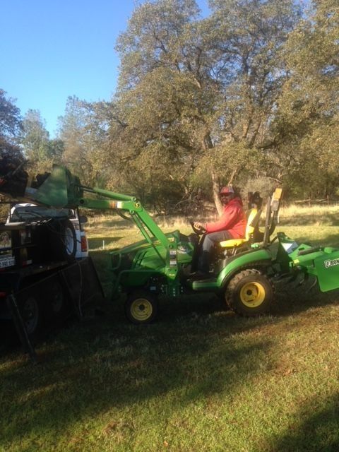 A person in a red jacket operates a green John Deere tractor with a front loader near a truck in a grassy field.