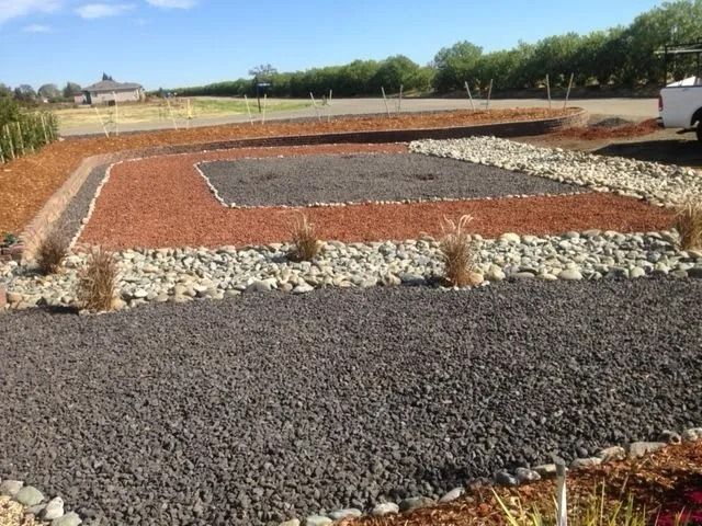 A landscaped yard featuring geometric patterns of red and grey gravel, river stones, and mulch under a sunny sky.