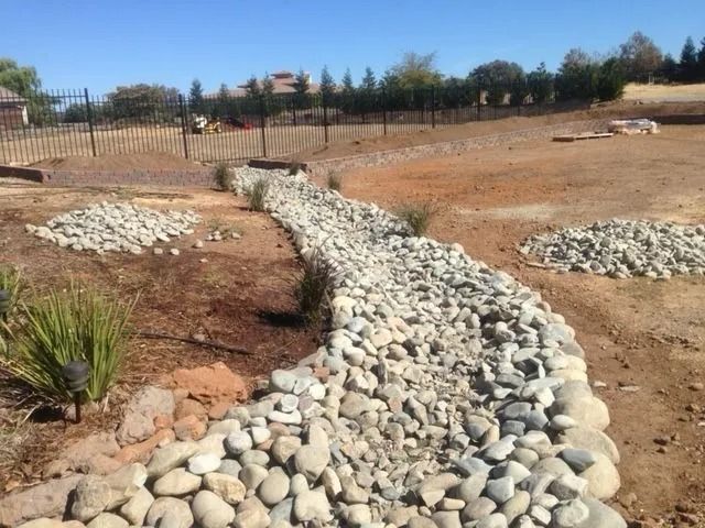 A dry creek bed made of river rocks winds through a landscape undergoing construction, with dirt areas and a metal fence.