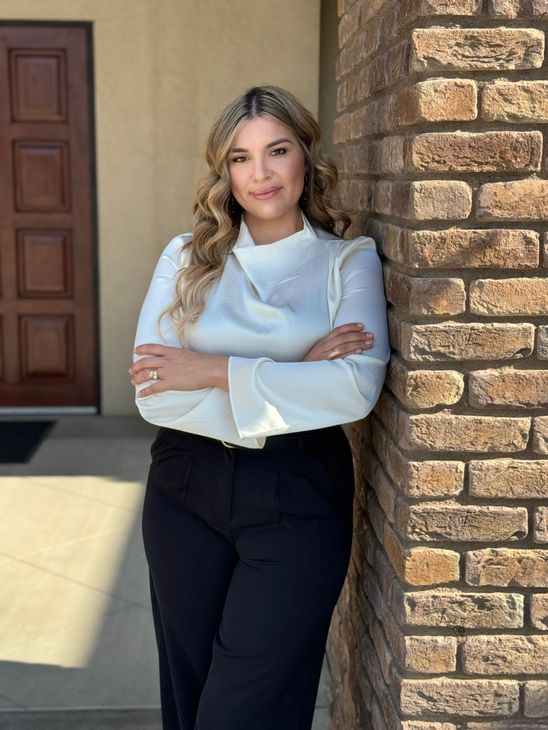 Woman in white blouse and black pants, leaning against a brick wall, arms crossed, smiling.