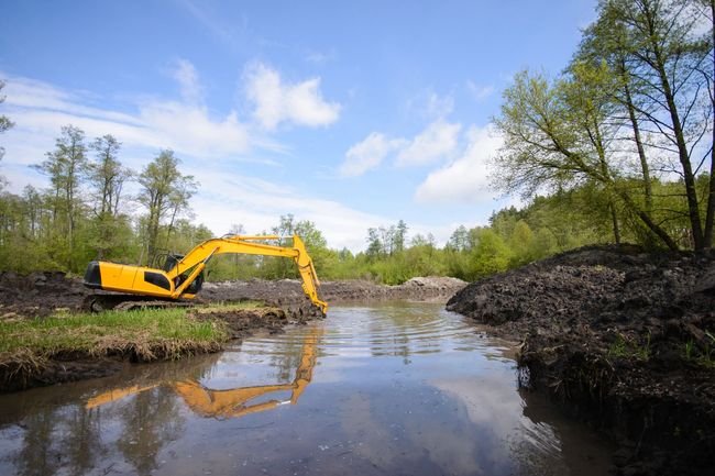 Yellow excavator dredging a murky river under a partly cloudy sky, near trees.