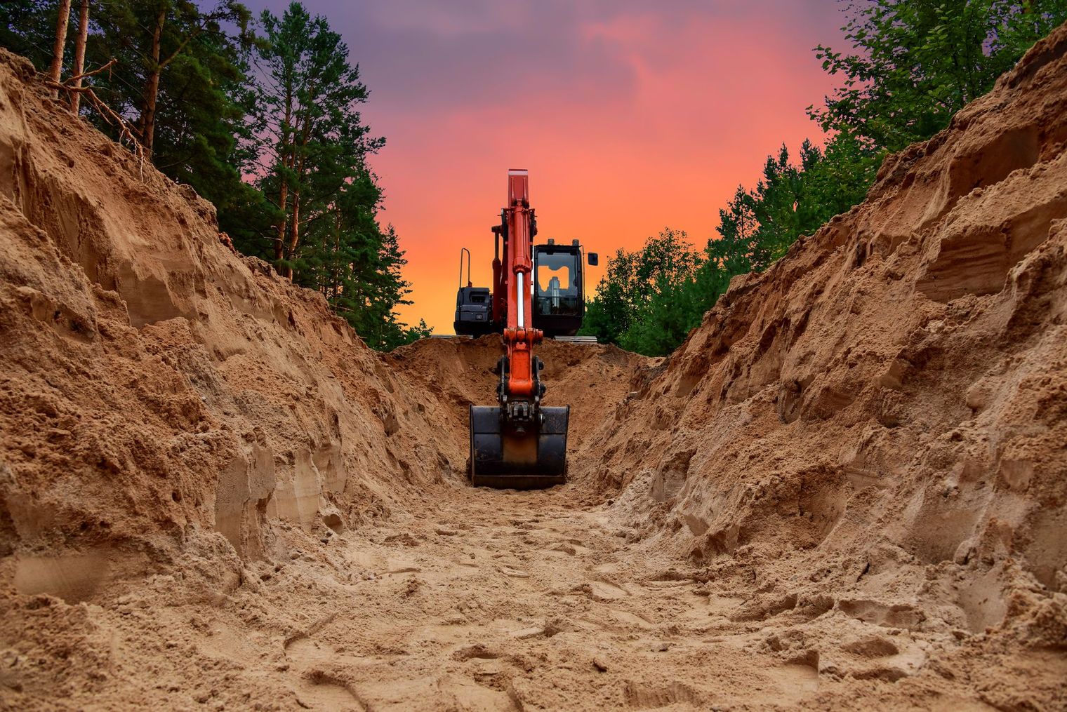 Excavator digging a trench in a sandy area, sunset sky in background.