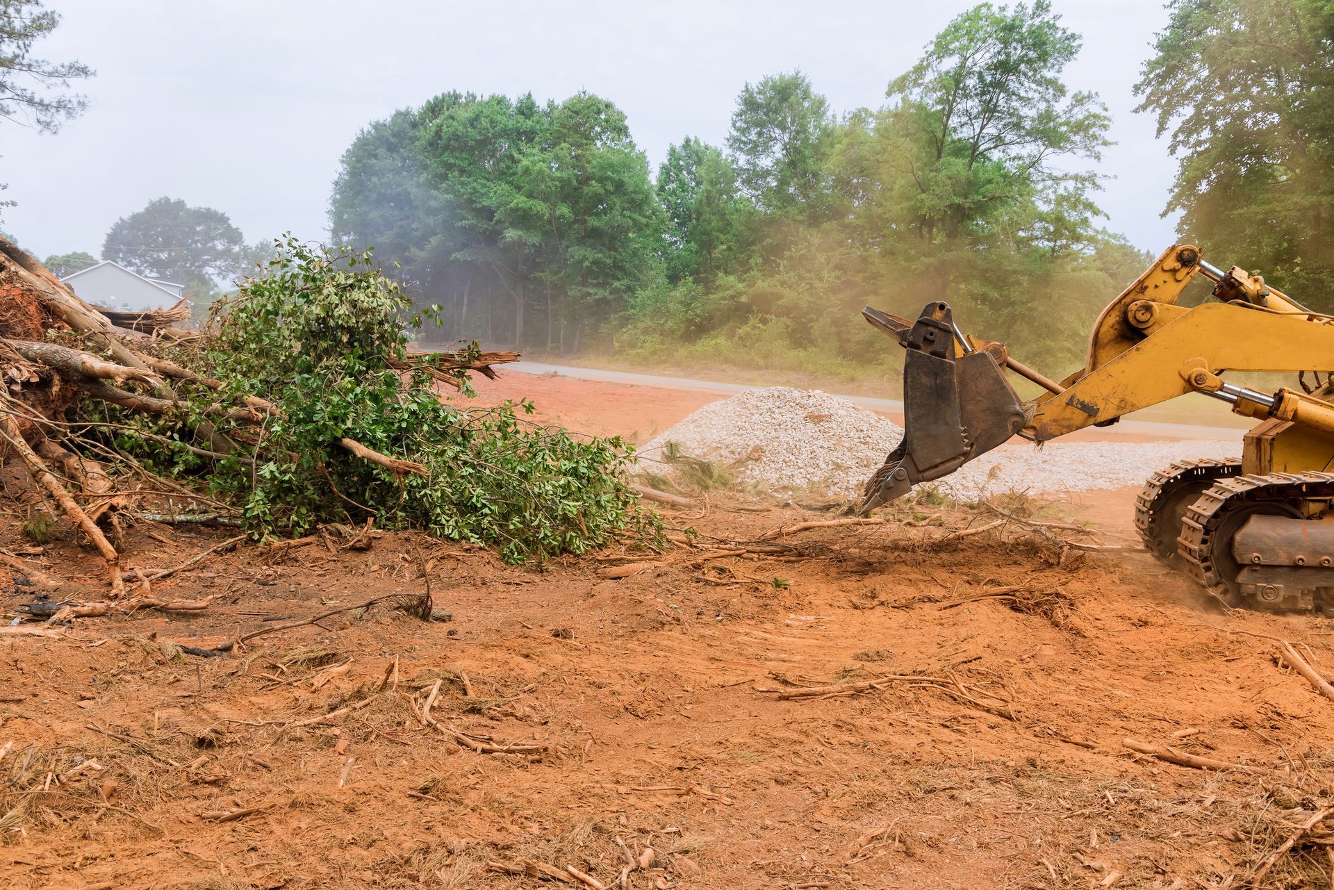 Yellow excavator clearing debris from a muddy construction site near a tree line.