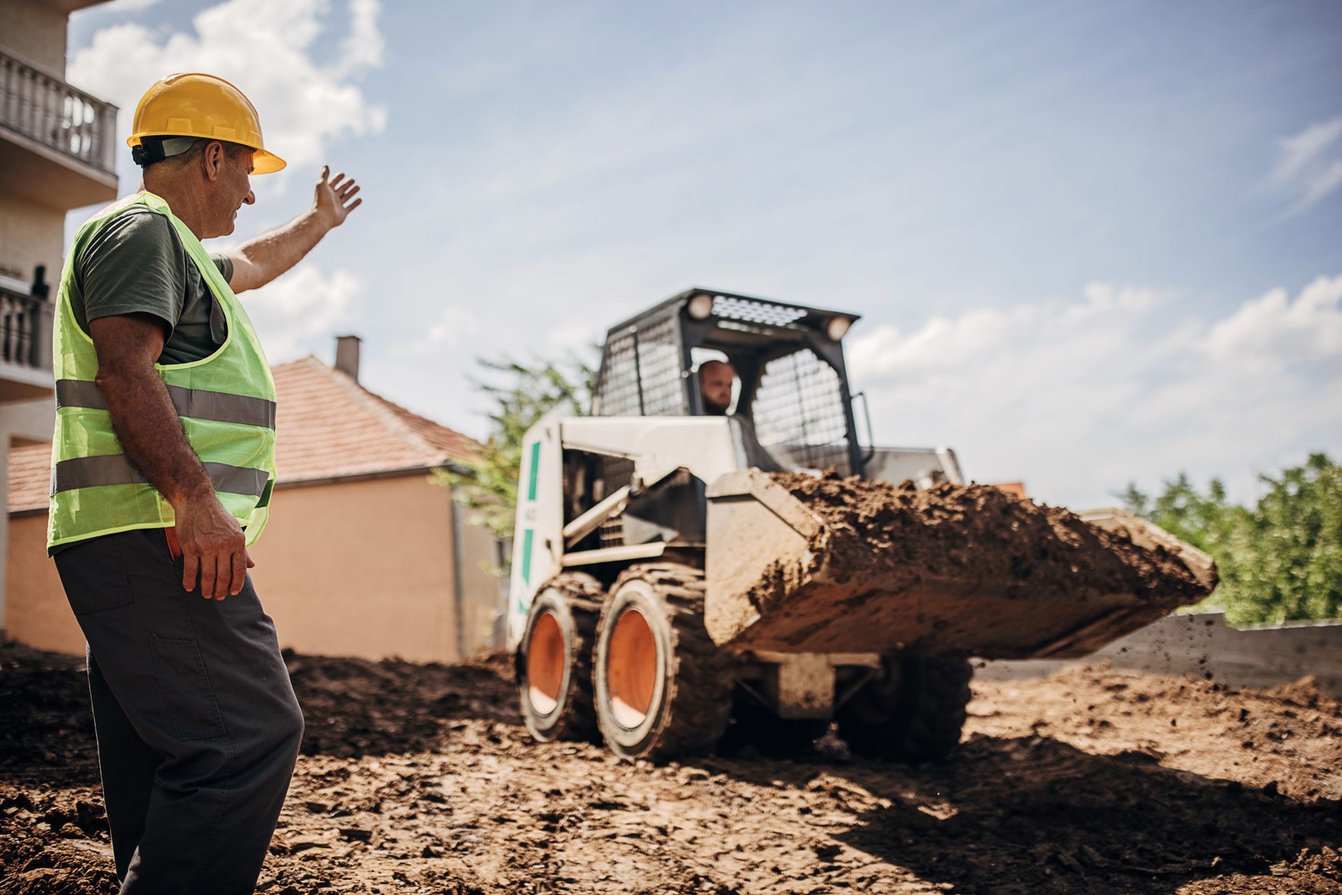 Man in safety vest directing a small loader carrying dirt on a construction site.