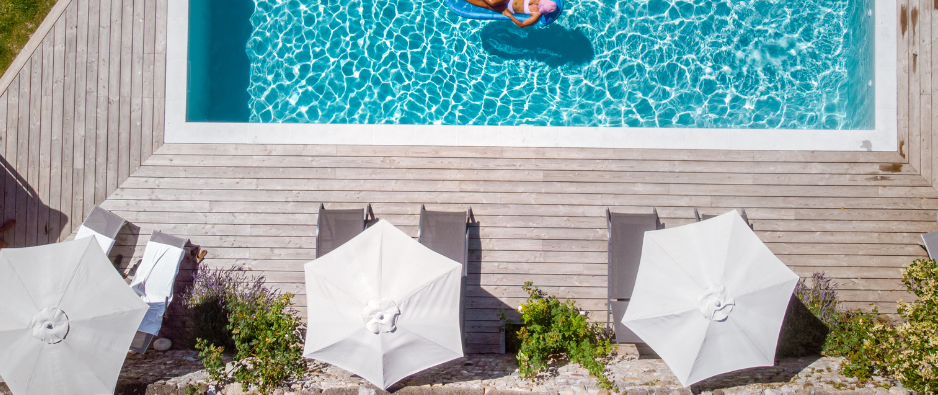 Overhead view of a pool with white umbrellas on a wooden deck and a swimmer in turquoise water